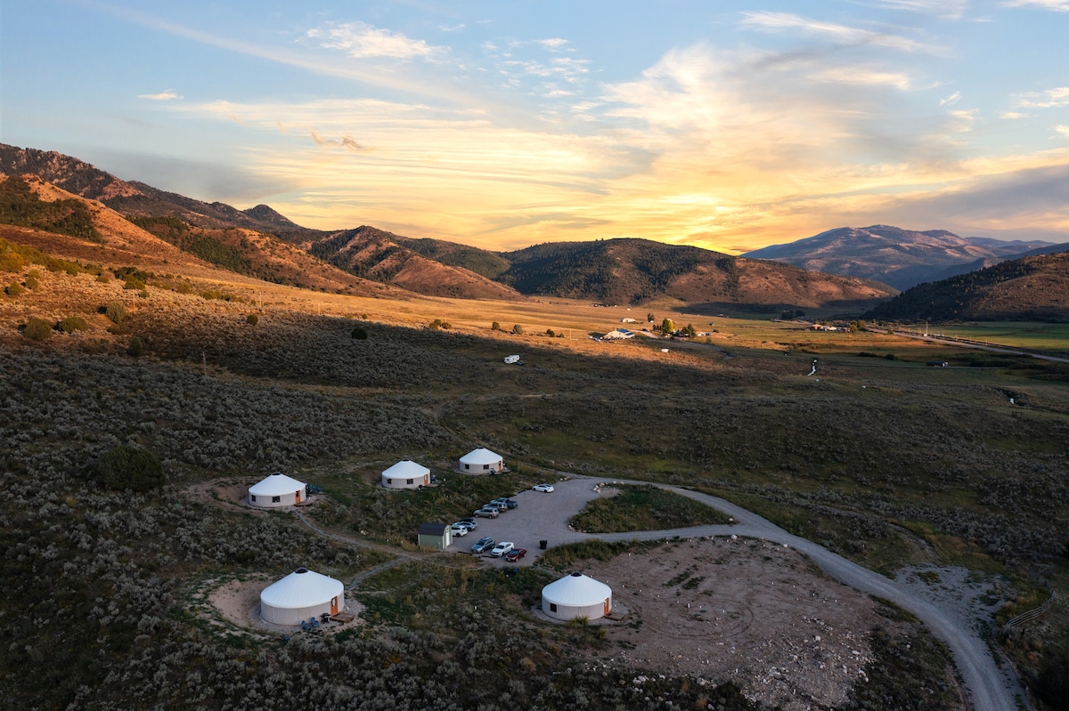 An aerial view captures five neatly arranged yurts on a hillside, surrounded by vast grassy fields and mountainous terrain. The landscape is illuminated by a warm sunset, highlighting the serenity of the retreat. A parking area is visible near the yurts, offering convenient access.