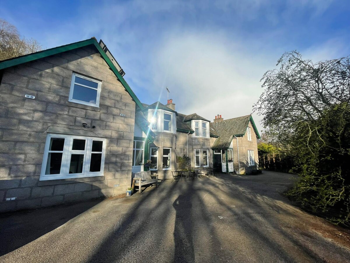 The exterior of a spacious bed and breakfast is framed by lush greenery, featuring a mix of stone and wooden architecture. Large windows reflect the daylight, while a welcoming entrance showcases a path leading to the front door. Shadows of surrounding trees add depth to the scene.