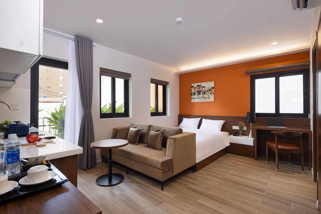 A well-lit room features a spacious bed against an accent wall. A comfortable sofa and a small dining table are visible. Windows allow natural light to enter, with curtains drawn to one side. A kitchenette is partly seen, equipped with essential appliances and utensils.