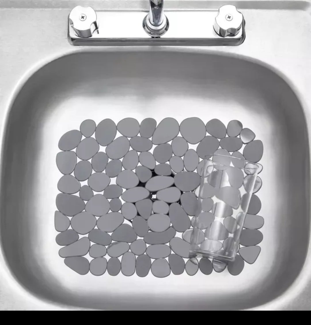 A modern stainless steel sink features a decorative arrangement of smooth, gray pebbles forming a pattern at the bottom. A transparent glass sits at the edge of the pebbles, indicating the functionality of the sink.