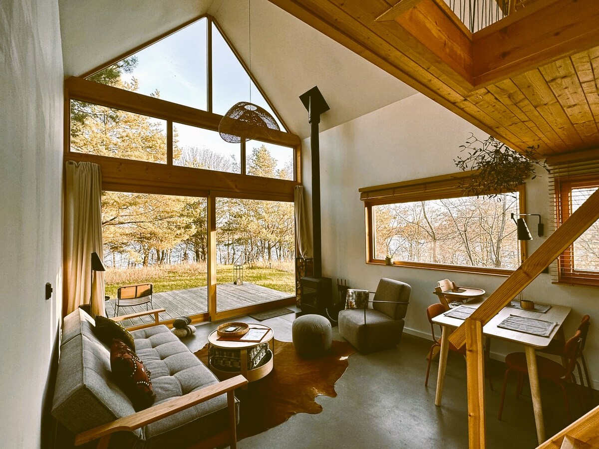 The living area features large front windows offering views of the natural surroundings. A spacious sofa and two comfortable chairs are arranged around a central coffee table. Wooden beams and warm tones create a cozy atmosphere, while a staircase leads to the upper level.