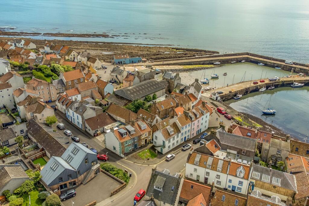 An aerial view captures a charming harbour village, showcasing a mix of quaint houses with varied rooflines. The coastline is visible, highlighting the working harbour with moored boats and rocky shores. Coastal pathways and residential streets create a serene atmosphere.