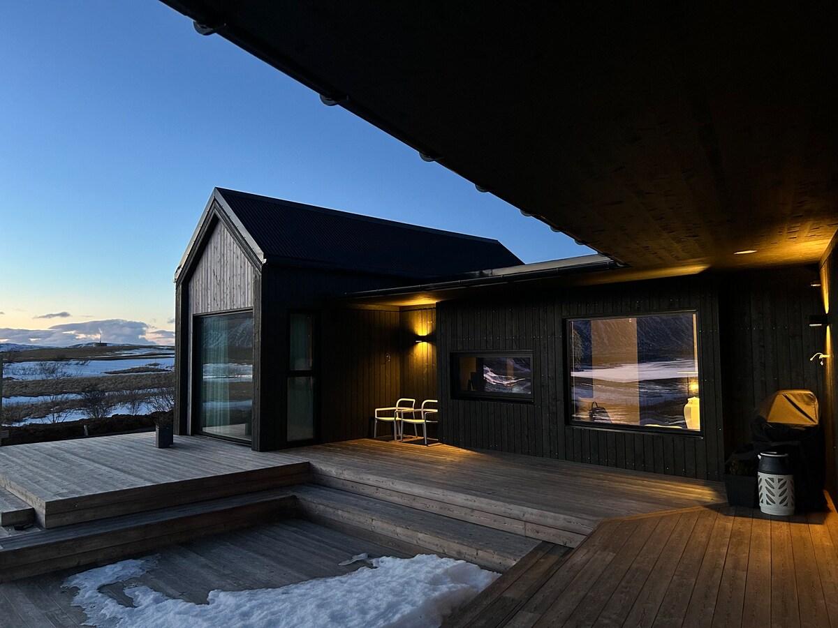 The exterior of the villa showcases a modern wooden facade, illuminated by soft outdoor lighting. A spacious wooden deck extends from the house, with two white chairs positioned for views of the surrounding landscape under a twilight sky.