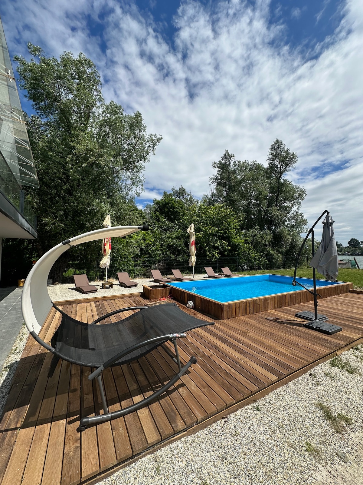 A wooden deck surrounds a private pool, offering loungers and umbrellas for shade. Lush greenery lines the background, providing a serene atmosphere. The sky is partly cloudy, and the pool's inviting blue water reflects the surrounding nature.