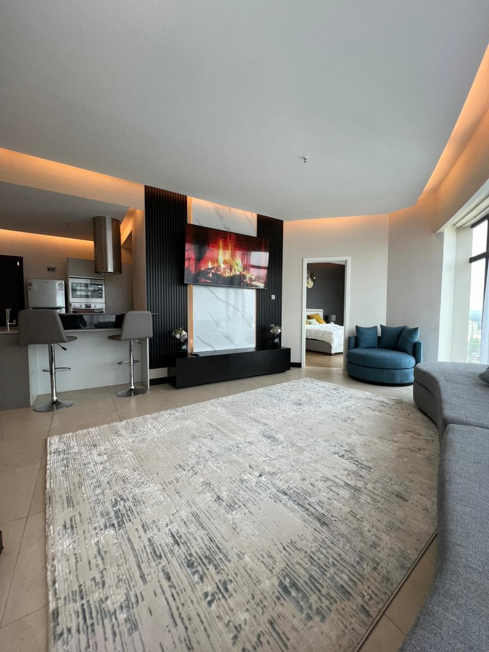 A modern living area showcases a plush gray sofa and a cozy circular blue chair. A large flat-screen television is mounted on a decorative wall, with a virtual fireplace display. The space is softly illuminated by ambient lighting, enhancing the contemporary aesthetic.
