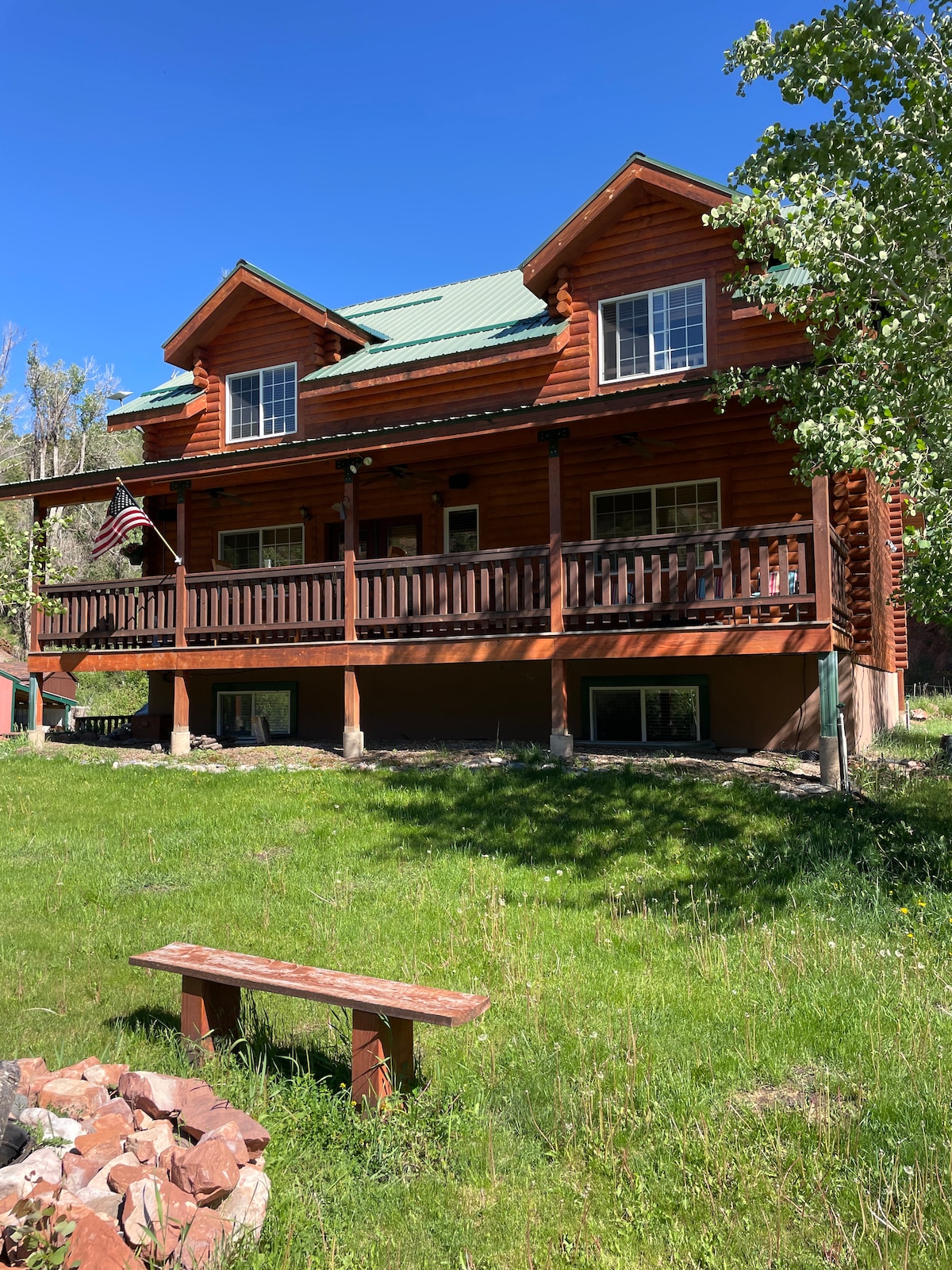 A spacious log cabin is featured, showcasing multiple levels with green metal roofing. A large deck spans the front, complemented by wooden railings and a bench in the foreground. Lush grass and trees surround the home, enhancing its natural setting.