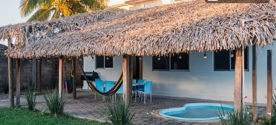 A thatched-roof terrace offers shaded seating with blue chairs and a hammock. A small plunge pool is visible on the side, surrounded by low-maintenance greenery. The exterior wall of the house is painted in soft tones, enhancing the overall tranquil environment.