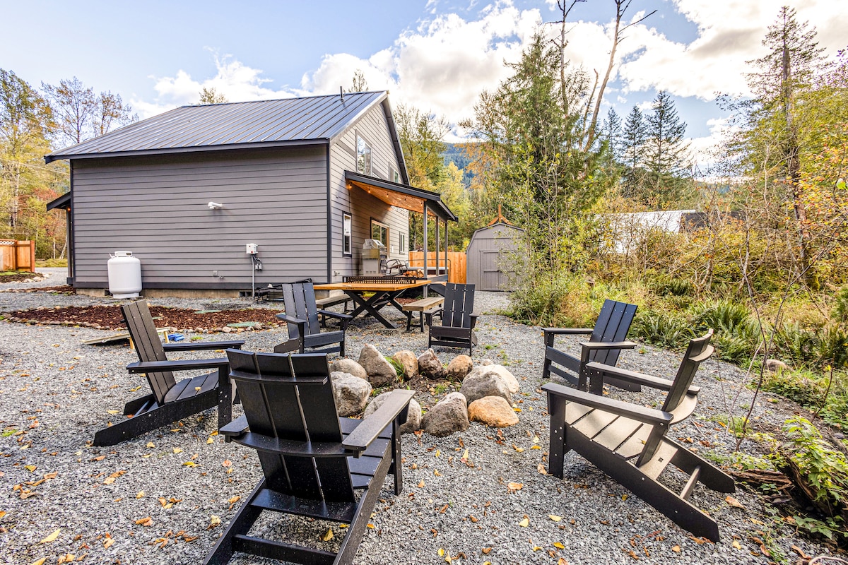 An outdoor fire pit area is surrounded by four Adirondack chairs, positioned on a gravel surface. A picnic table and additional seating are visible under a covered patio attached to the cabin. Lush greenery and trees frame the background, enhancing the serene atmosphere.