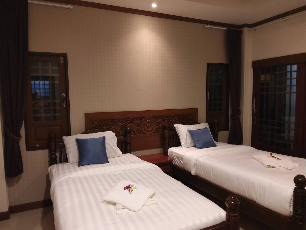 The sleeping area features two wooden beds adorned with white linens and plush pillows. Decorative wall elements and large windows allow natural light to fill the room, while dark curtains provide options for privacy and light control.