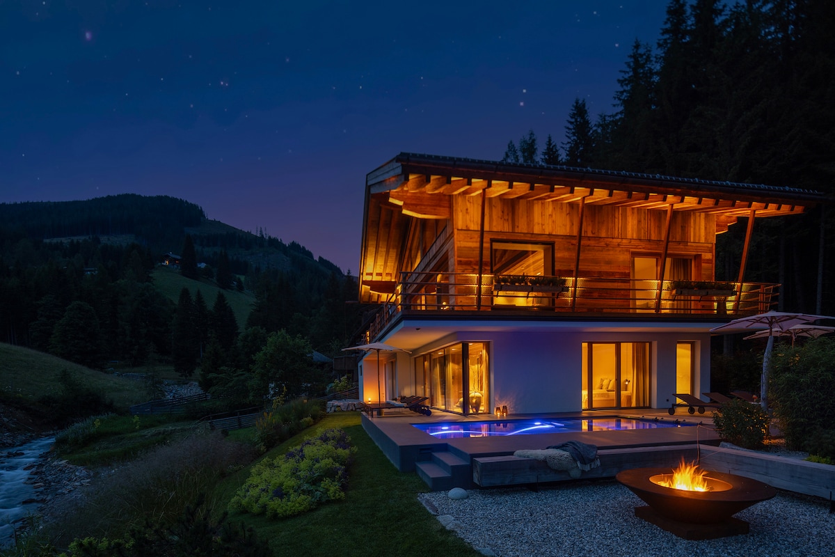 The chalet is illuminated at night, showcasing its modern architecture. An outdoor pool reflects warm light, while a cozy fire pit rests nearby on the terrace. Surrounding greenery and mountain views create a serene atmosphere.