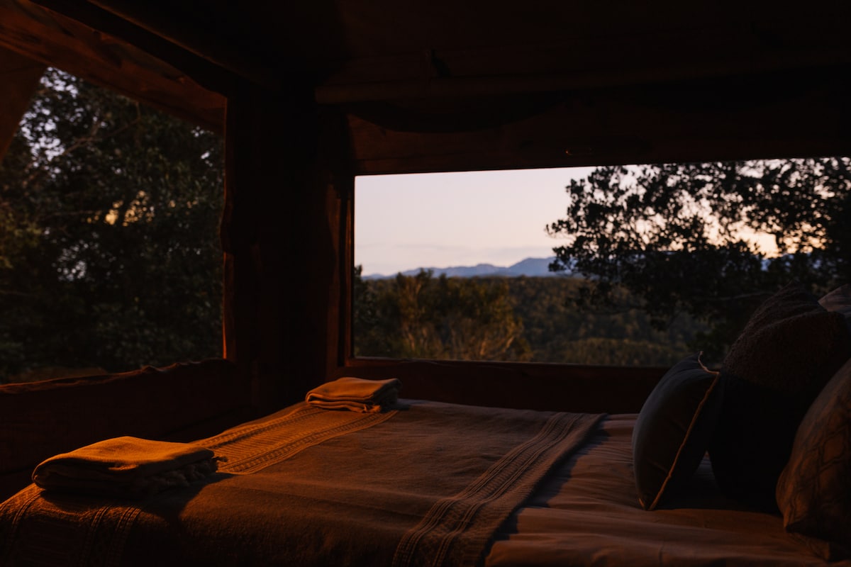 A serene sleeping area features a spacious bed adorned with neutral-toned linens. Large windows provide a glimpse of the surrounding forest and distant mountains, allowing natural light to gently illuminate the space. Soft towels are placed thoughtfully on the bed.