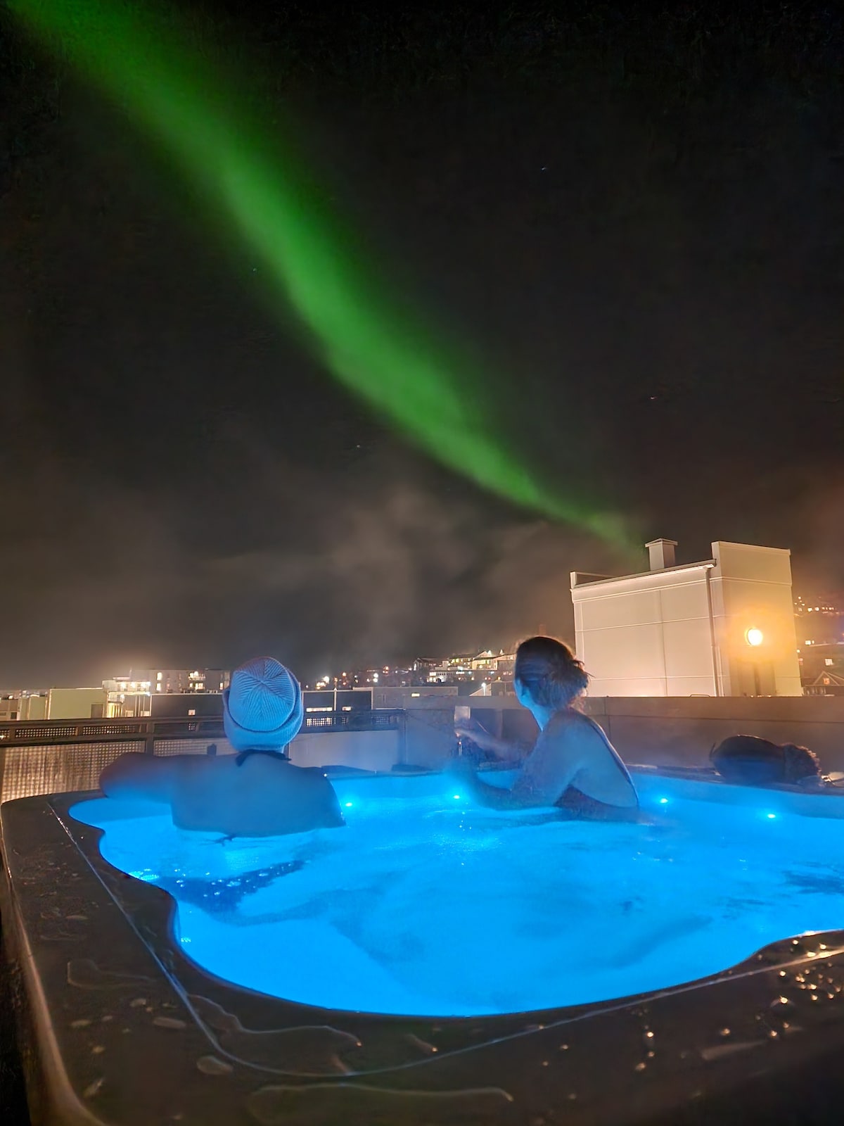 Two individuals are observed relaxing in a spacious jacuzzi, surrounded by softly glowing blue water. The northern lights illuminate the night sky above, casting a vibrant green hue, while city lights twinkle in the background, enhancing the serene atmosphere.