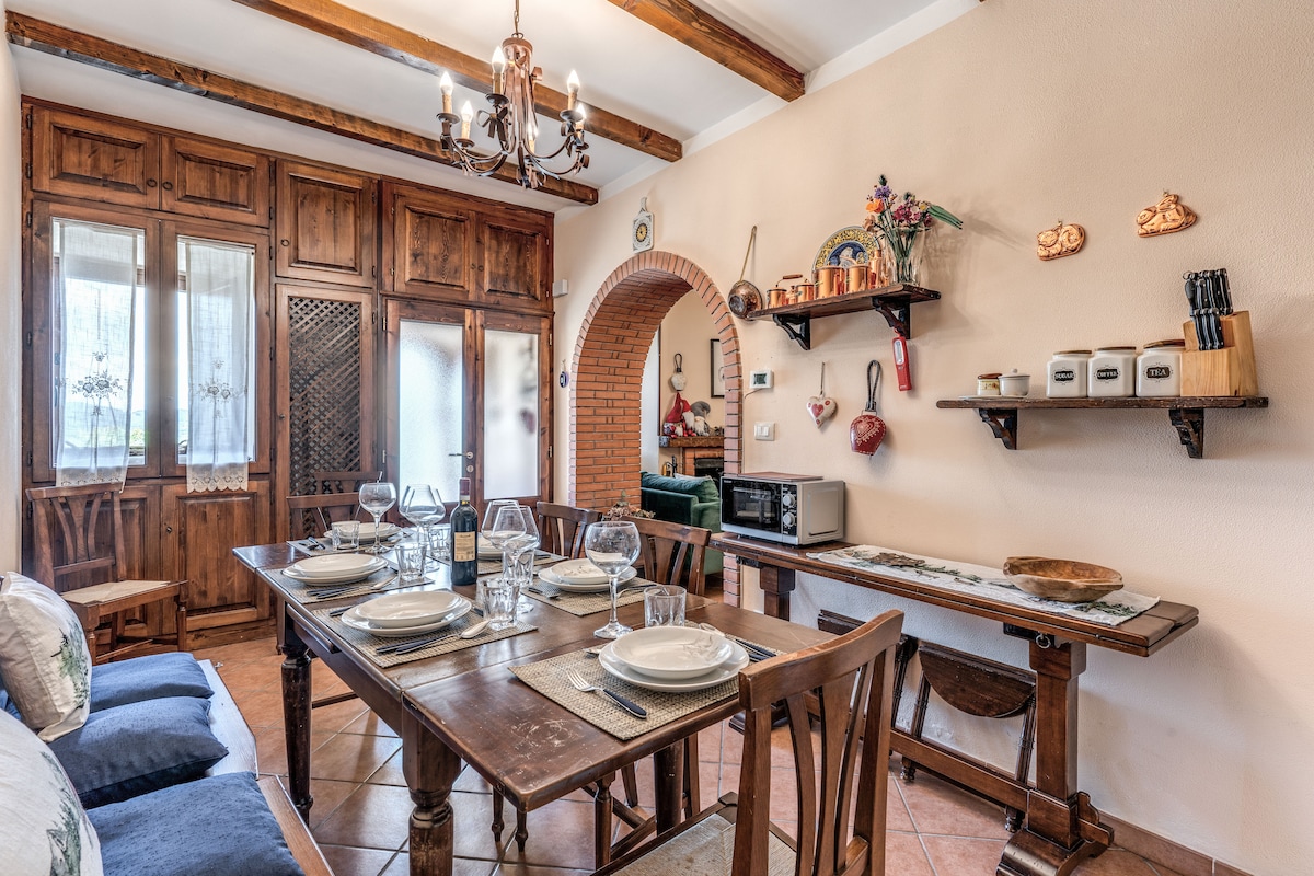 A warm dining area is presented with a wooden table set for six, complete with glassware and placemats. Surrounding wooden cabinetry offers storage, while a rustic shelf showcases kitchen items. The space is enhanced by exposed wooden beams and a charming chandelier.