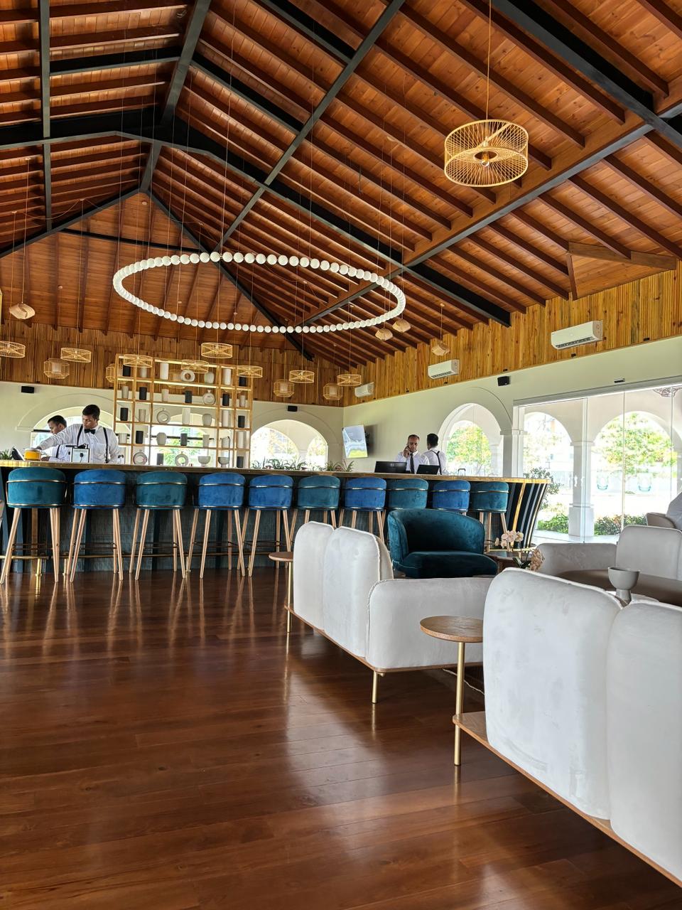 A spacious bar area features high-backed blue chairs surrounding a sleek bar. The wooden ceiling with exposed beams creates a warm atmosphere, while large arched windows invite natural light. Stylish seating arrangements are present, complemented by a modern chandelier that adds a touch of elegance.