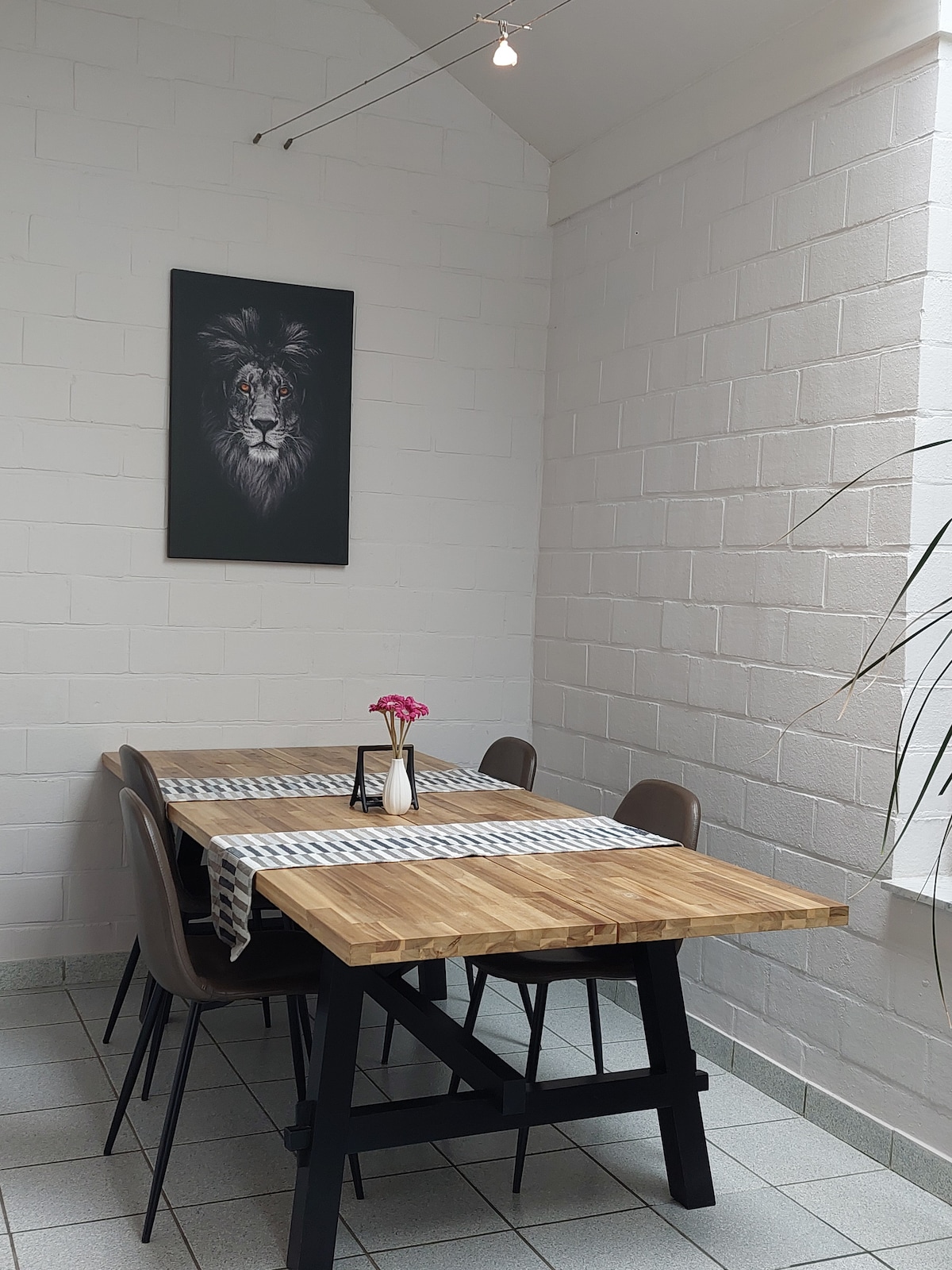 A dining area is featured with a wooden table accompanied by four chairs. A decorative centerpiece with a flower is placed on the table. A striking artwork of a lion adorns the wall, with soft lighting enhancing the space's ambiance.