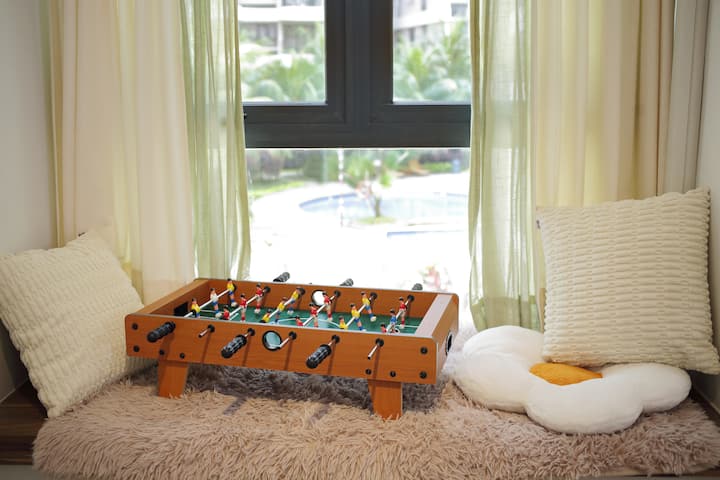 Un acogedor rincón con una pequeña mesa de futbolín sobre una alfombra esponjosa, rodeada de cojines suaves, que ofrece un lugar lúdico para relajarse con vistas a la vegetación y a la piscina exterior.