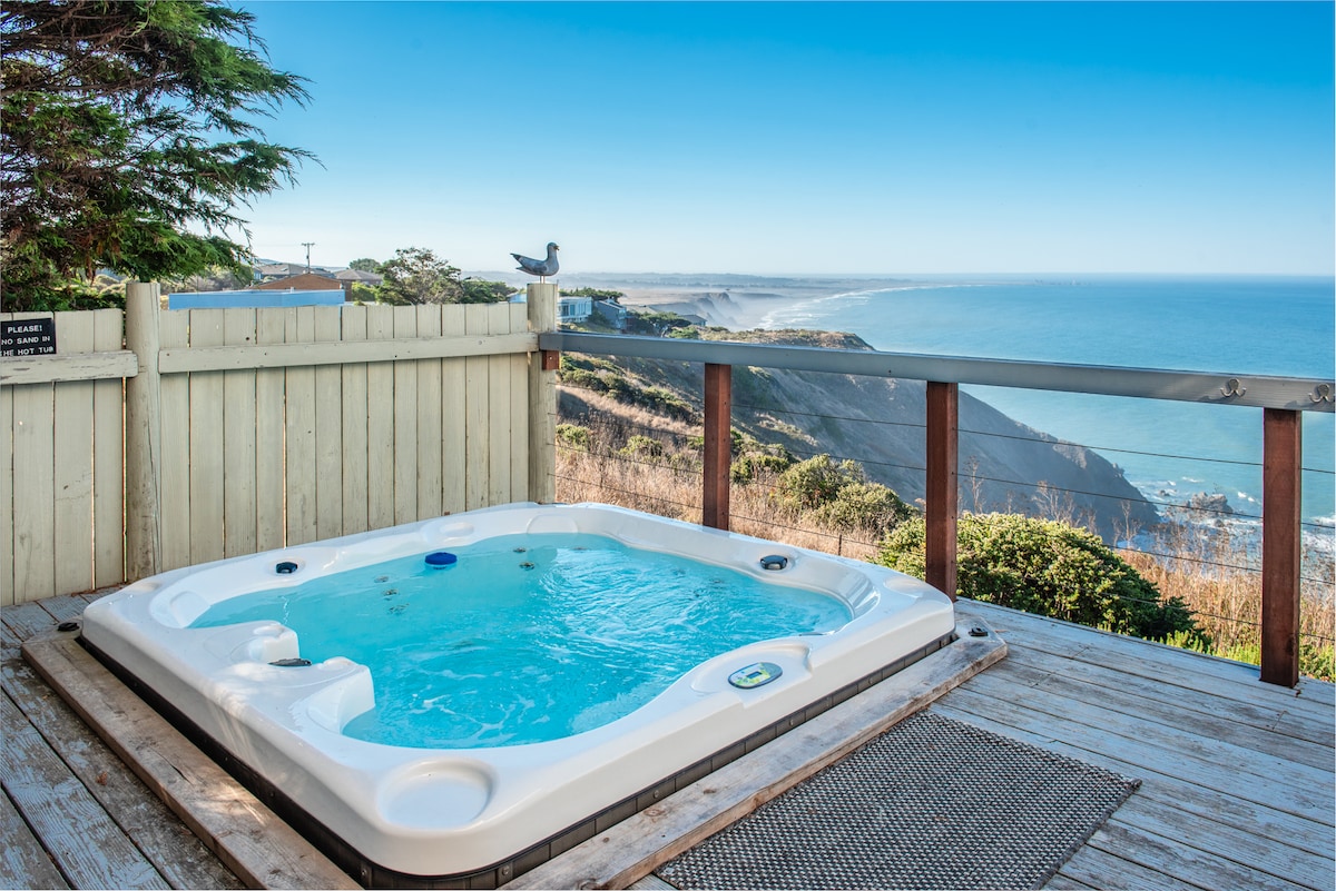 A sunken hot tub is positioned on a wooden deck, offering expansive views of the ocean and coastline. The clear water reflects the sky, while a railing encloses the area. Coastal trees provide a natural backdrop to the serene setting.