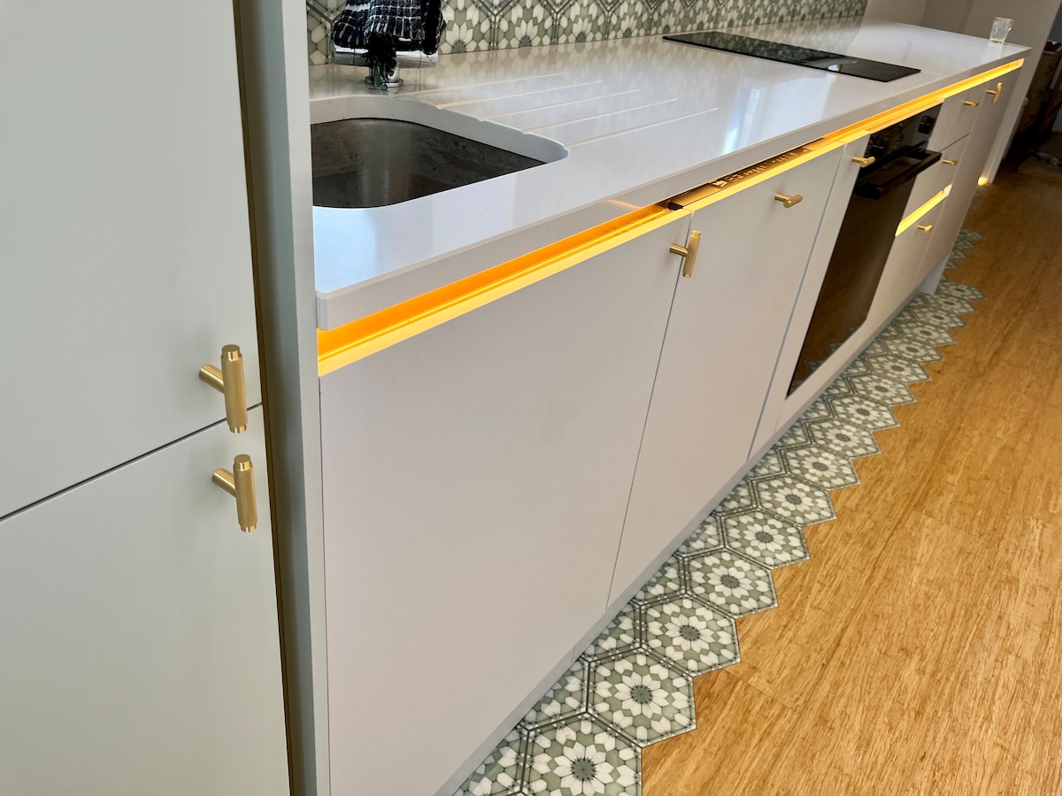 A contemporary kitchen features sleek cabinetry with gold hardware and underlit surfaces. Dual sinks are visible, along with integrated appliances, providing a modern aesthetic. The patterned tile flooring adds texture, harmonizing with the overall design.