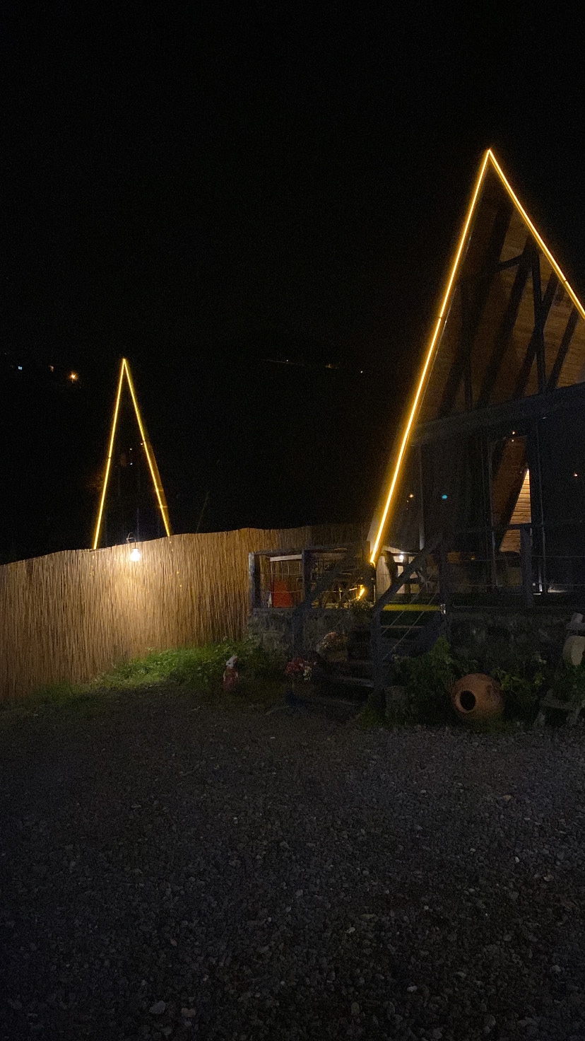 Two charming A-frame bungalows are illuminated by soft, ambient lighting against a dark night sky. A rustic wooden fence and gravel pathway lead to the entrance, while subtle landscaping features complement the serene environment.