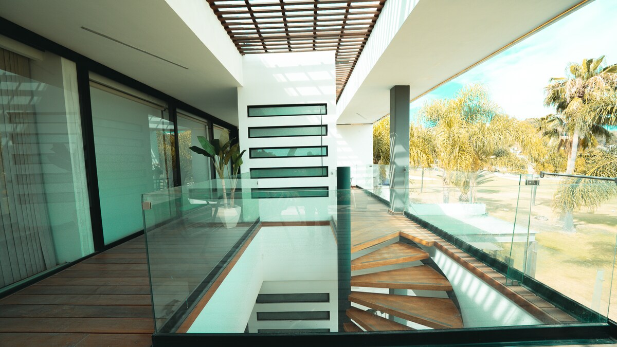 A modern open space is showcased, featuring wooden flooring and glass railings. Large windows allow natural light to fill the area, offering views of palm trees outside. A staircase with wooden steps descends to a lower level, emphasizing the spacious architecture.