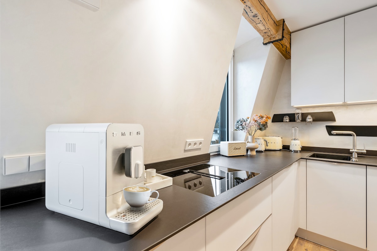 A modern kitchen is showcased with sleek cabinetry and a dark countertop. An espresso machine is prominently placed beside a small cup, inviting guests to enjoy a warm beverage. Natural light streams through a window, illuminating the stylish decor and kitchen appliances.
