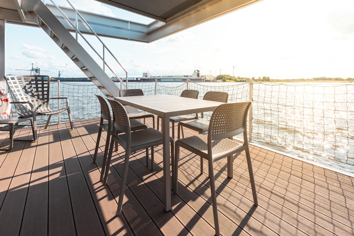 A spacious outdoor terrace features a large table surrounded by six chairs, providing a comfortable area for dining or relaxation. A sleek staircase leads to the upper deck, where views of the water and surroundings can be enjoyed.