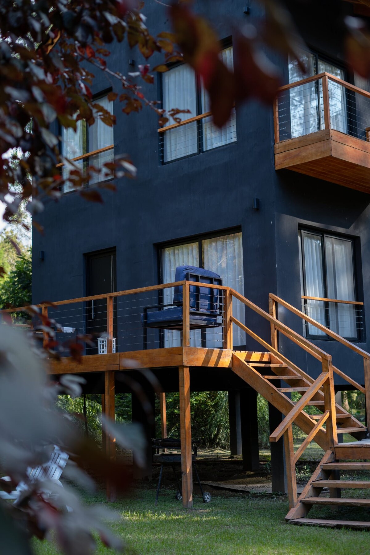 A modern cabin exterior showcases a dark facade and wooden deck. Steps lead to the entrance, where outdoor seating is positioned on the deck. Large windows allow natural light to enter the interior space, surrounded by lush greenery.