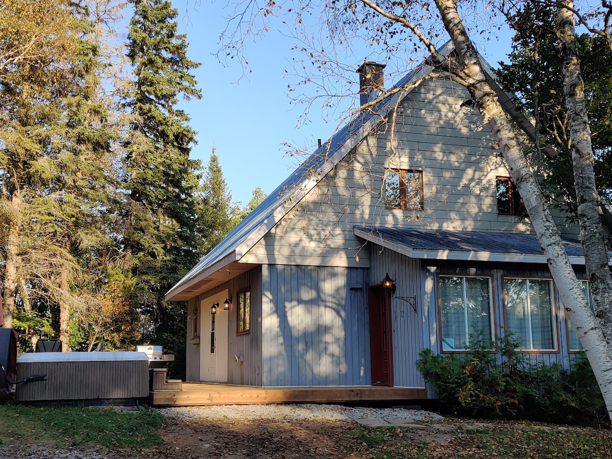The chalet features a two-story wooden exterior with large windows allowing natural light in. A spacious deck leads to a private spa area, and surrounding trees provide a sense of privacy. The welcoming front door is complemented by outdoor lighting.