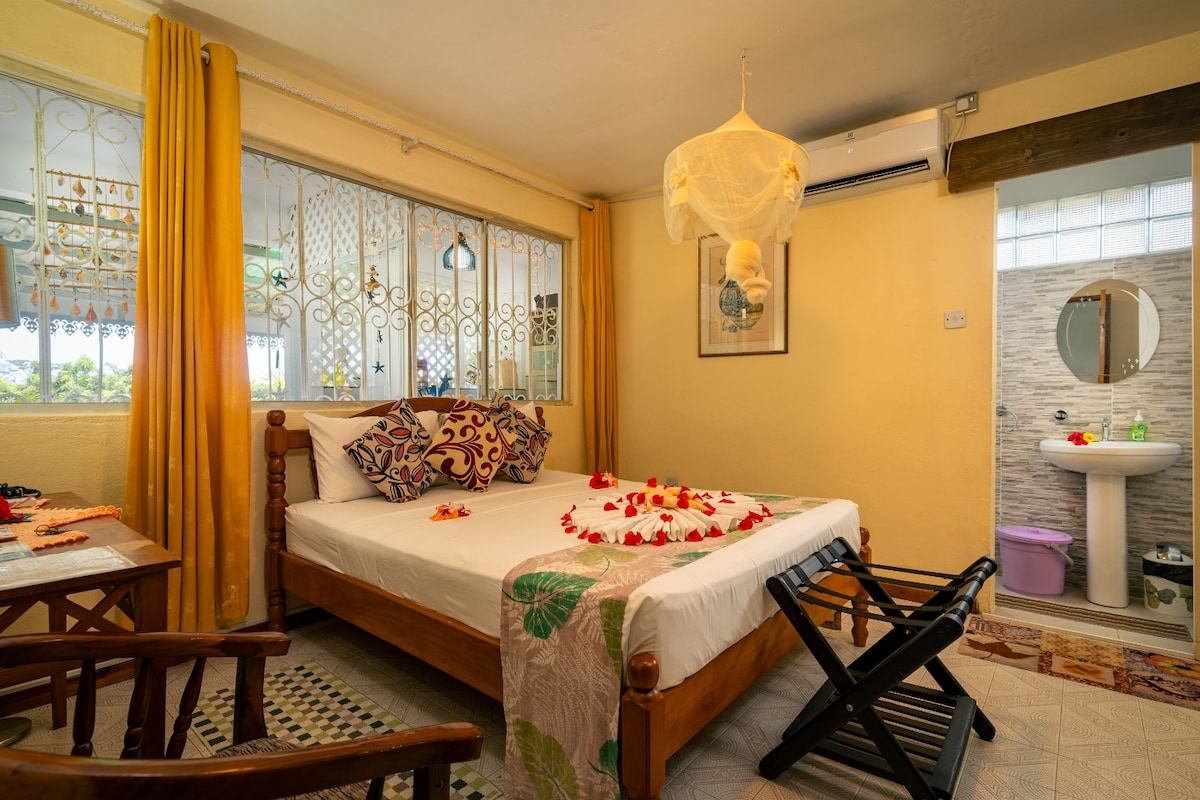 The room features a queensize bed adorned with flower petals, complemented by decorative pillows. A wicker chair and a folding stool are positioned nearby. The en suite bathroom includes a round mirror and essential amenities. A bright window allows natural light to fill the space.