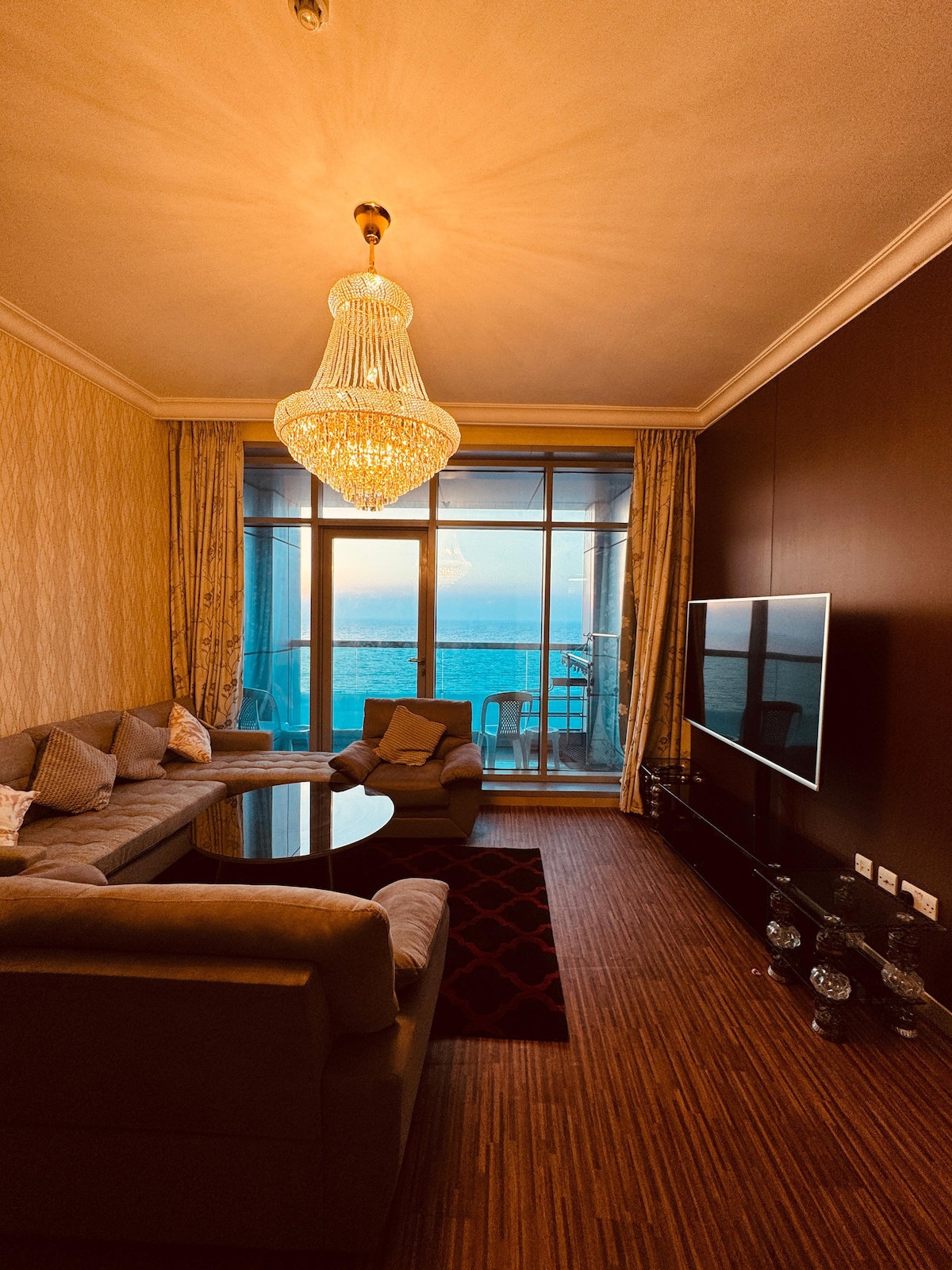 A spacious living area is highlighted by a crystal chandelier, which hangs above a stylish seating arrangement. Large glass doors provide a panoramic view of the Arabian Sea, with light streaming in and softly illuminating the room's warm decor.