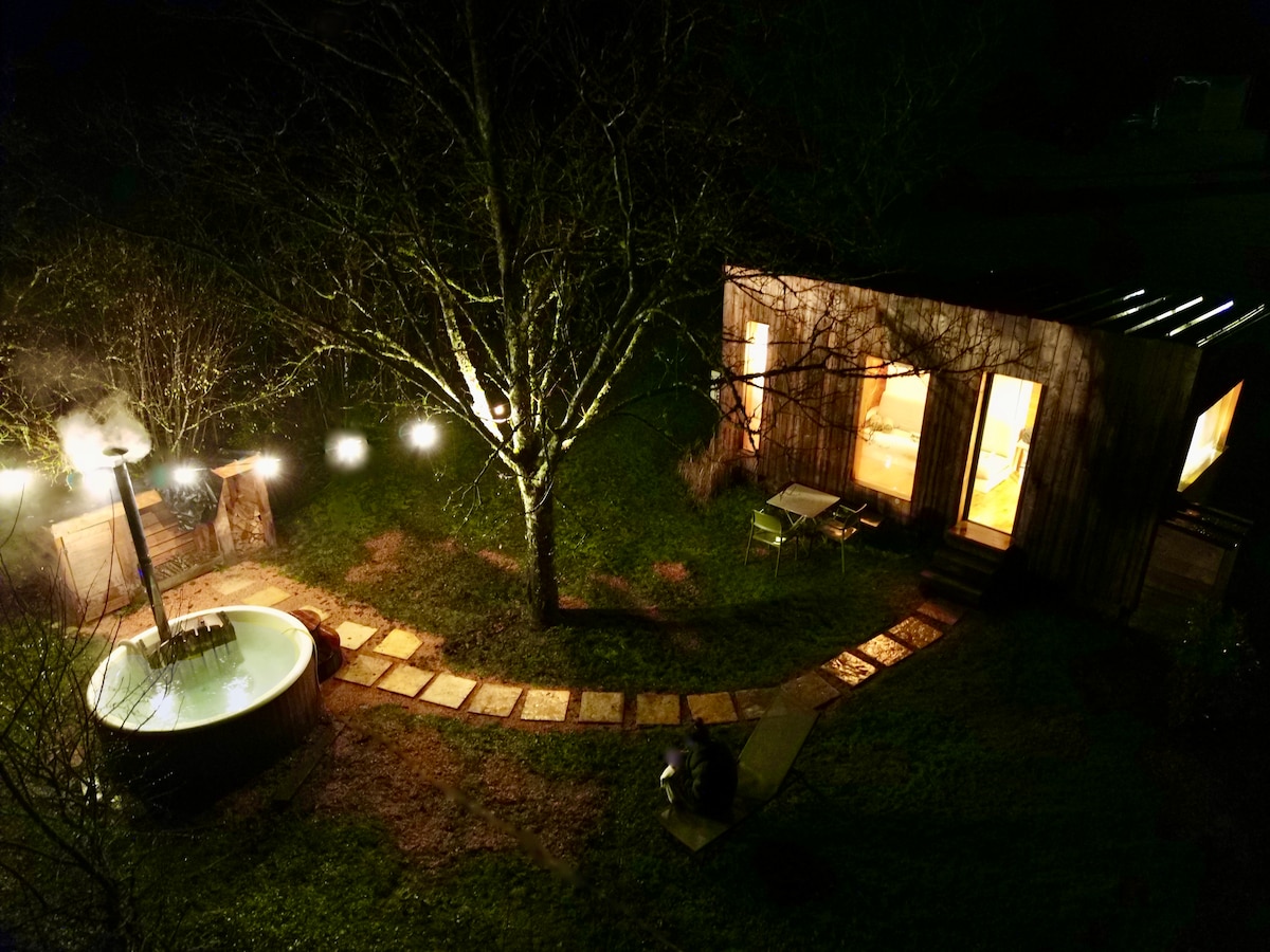 A wooden cabin sits in a serene outdoor setting, with illuminated windows and a warm glow emanating from within. A circular wood-fired hot tub is placed nearby, surrounded by a stone path and tree branches, enhancing the tranquil atmosphere of the night.
