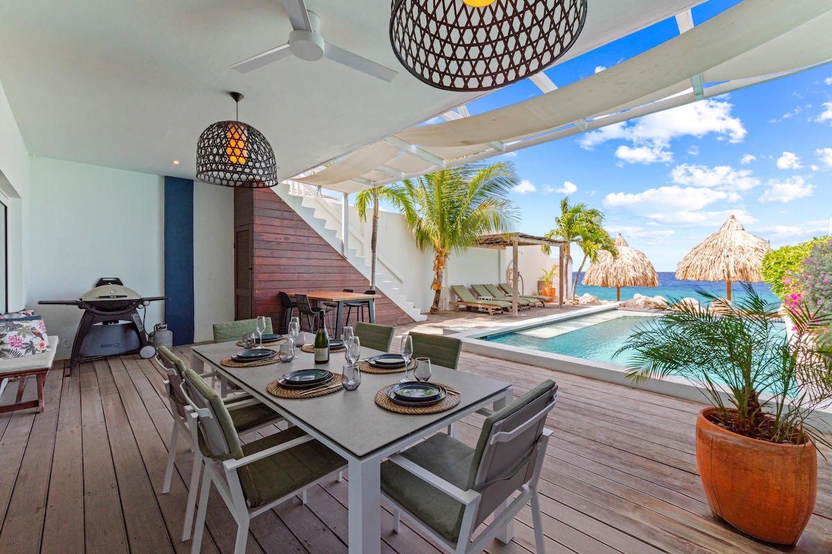 An outdoor dining area is presented with a large table and multiple chairs, accompanied by stylish light fixtures. A pool is visible in the background, alongside sun loungers and palapa-style cabanas, framed by lush greenery and a clear blue sky.