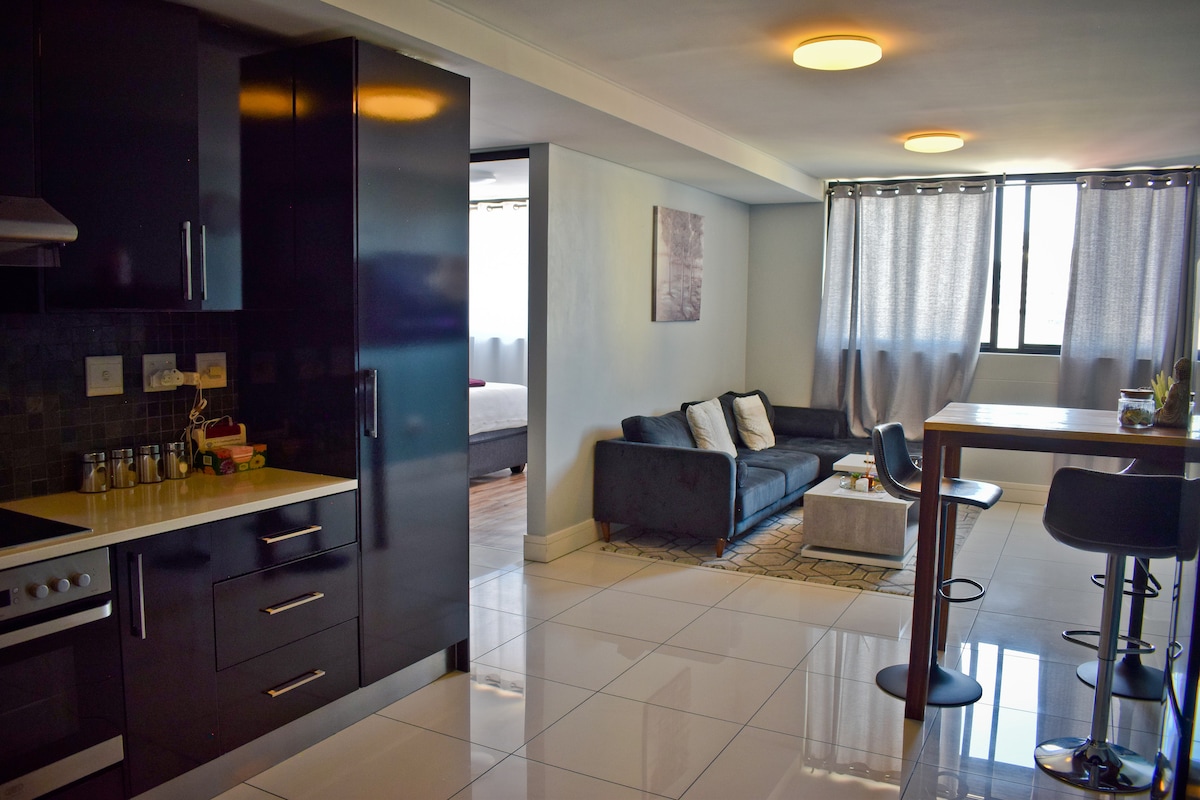 A modern open-plan living area features a sleek kitchen with dark cabinetry and a white countertop. A cozy sofa sits adjacent to a high dining table with four stools, while light is filtered through sheer curtains at the window.