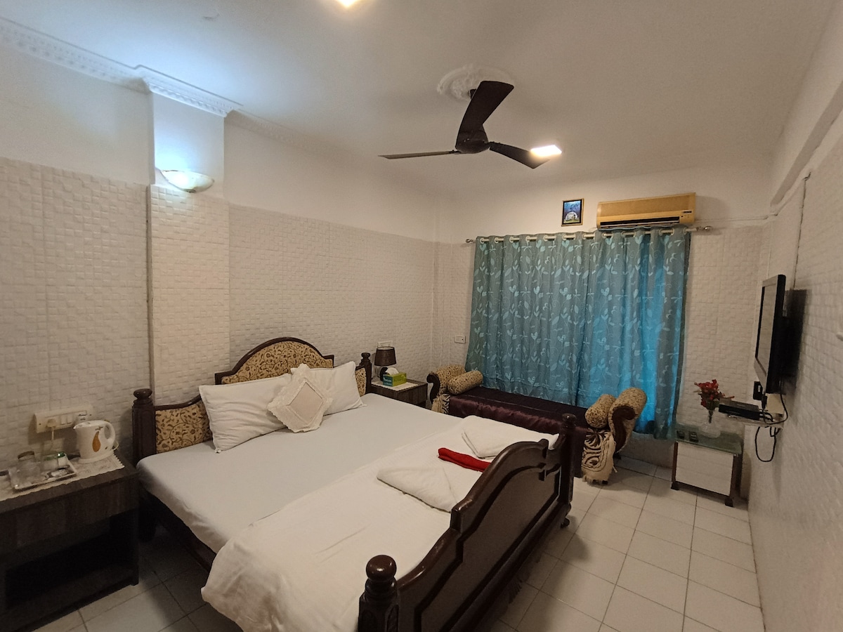 A spacious room features a large bed with crisp linens and decorative pillows. Two armchairs are positioned near a window adorned with light blue curtains. A mounted television and a small table are visible, along with a ceiling fan providing ventilation.