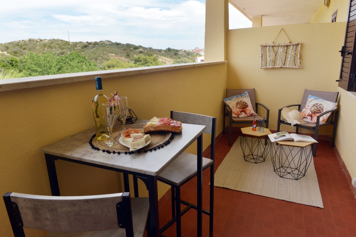 A cozy balcony area features a small dining table set with a bottle of wine, glasses, and a plate of food. Two comfortable chairs with decorative cushions complement a textured rug, while the surrounding landscape offers a view of greenery.