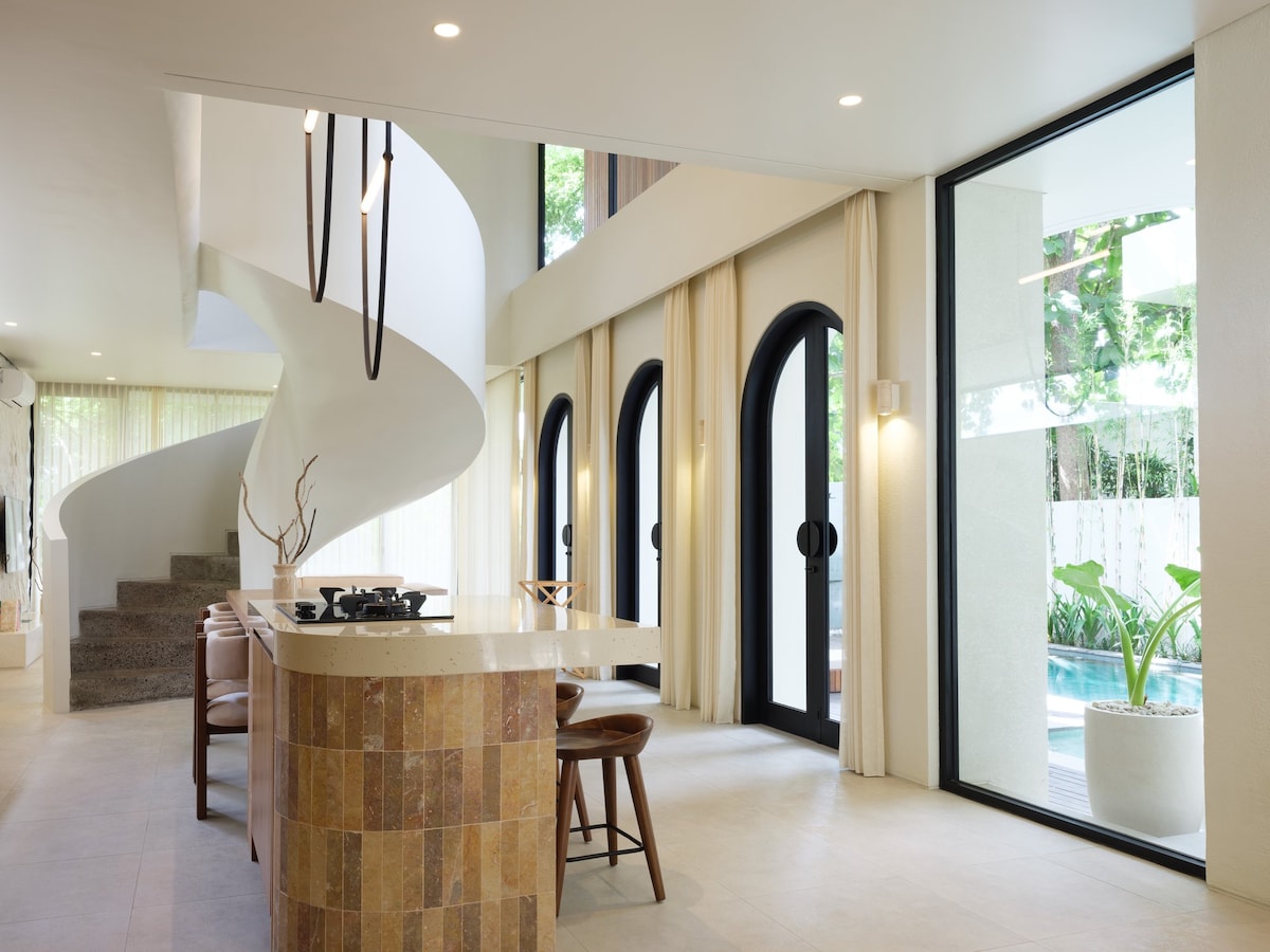 An open-plan living area features a modern kitchen island with natural stone accents and wooden seating. Large glass doors frame views of the pool outside, while a sweeping staircase with a sculptural design adds elegance to the space. Natural light floods the room through multiple windows.