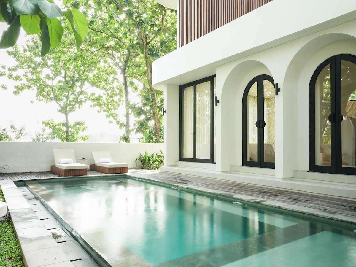 A private pool lies on a wooden terrace, surrounded by lush greenery. Two lounge chairs are positioned poolside, offering a serene spot for relaxation. Expansive glass doors provide a view of the bright interior, seamlessly connecting outdoor and indoor spaces.