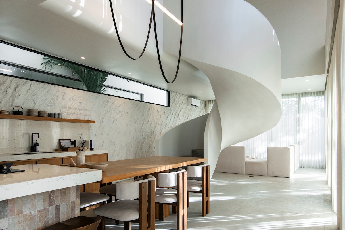 An open-concept interior showcases a modern dining area with a wooden table and upholstered chairs, accompanied by a sleek kitchen. A striking spiral staircase ascends to the upper level, creating a dynamic focal point within the spacious living area bathed in natural light.
