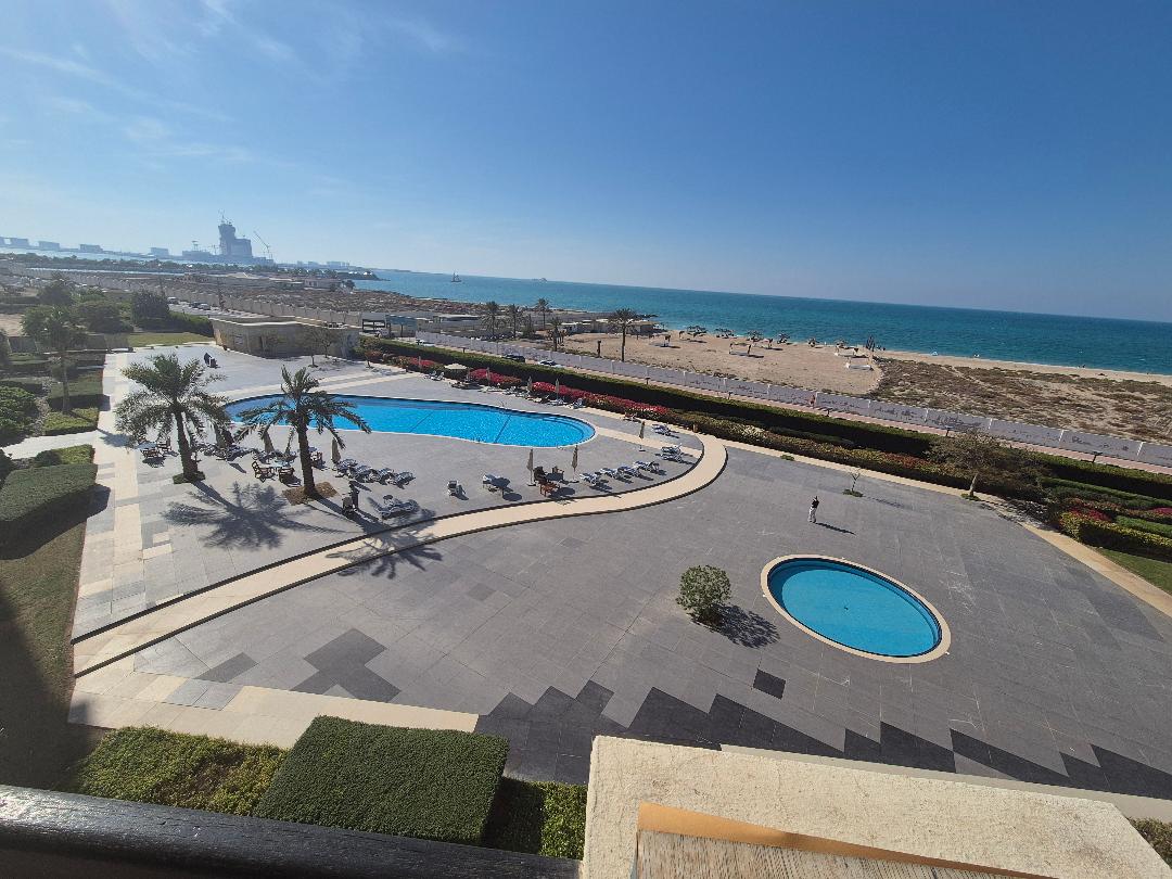 A view overlooking a large outdoor pool area features smooth stone flooring. Sun loungers are arranged around the pools, with palm trees providing shade. In the background, the beach meets the sea, contrasting with the greenery of the landscaped grounds.