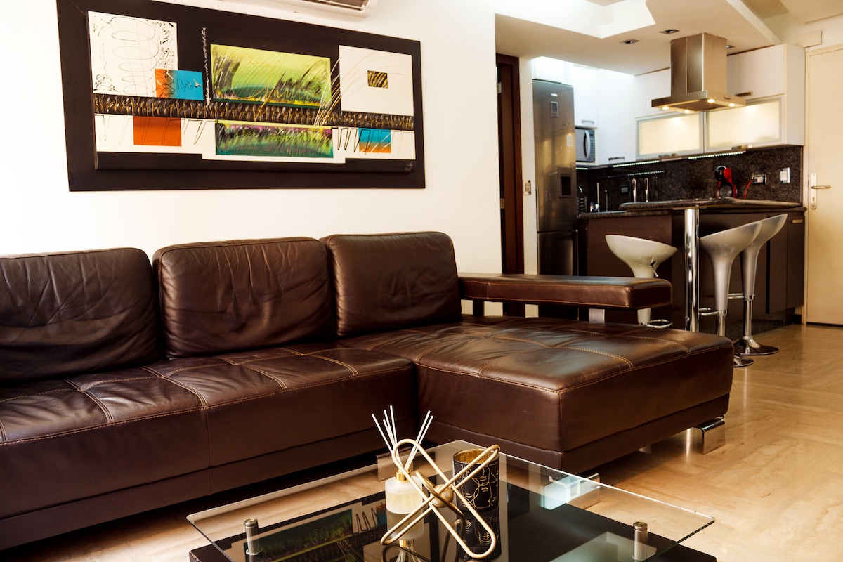 A modern living area features a large brown leather sectional sofa complemented by a sleek glass coffee table. A colorful artwork hangs on the wall, and the open kitchen is visible in the background, equipped with contemporary appliances and bar seating.