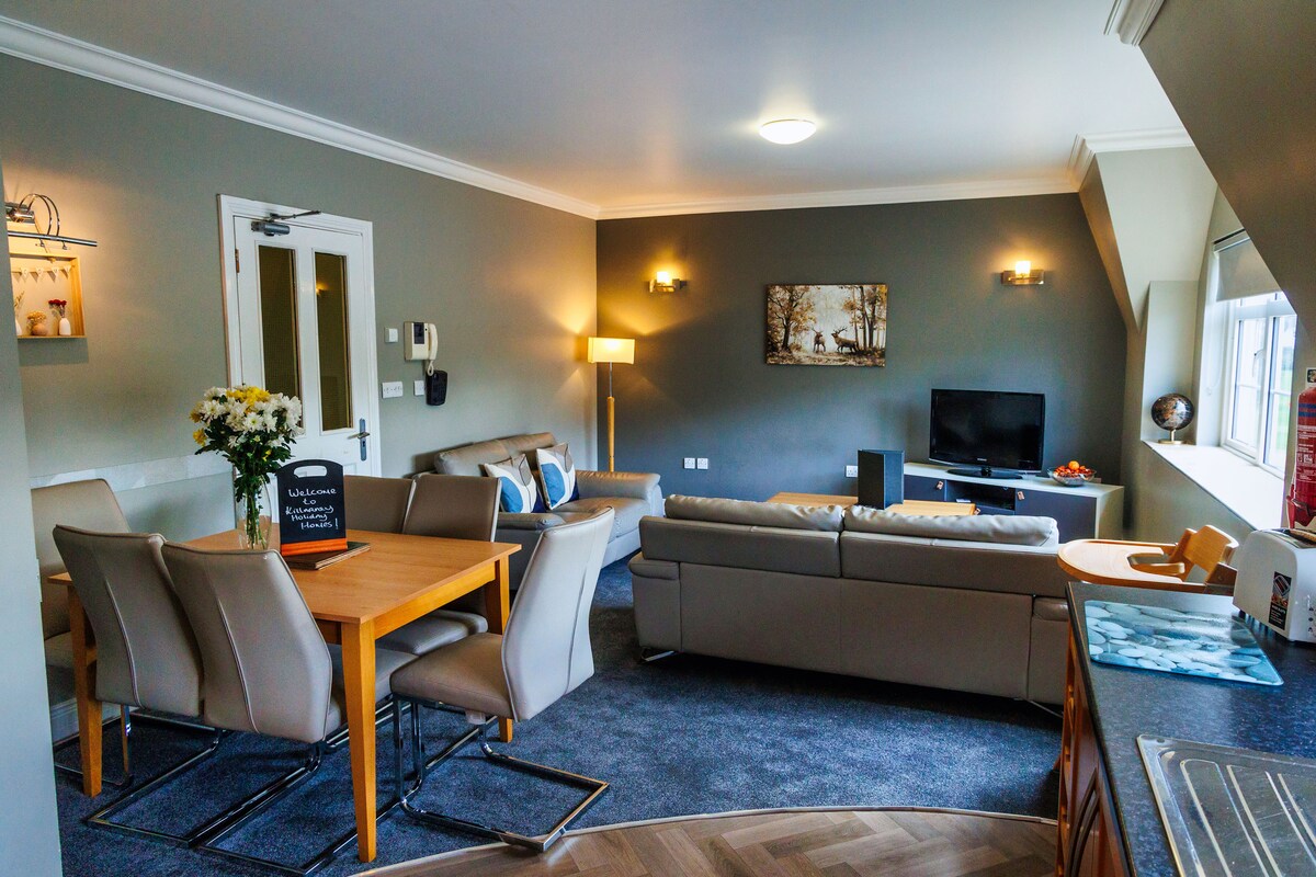 A modern living area is presented with a soft grey color scheme. A comfortable seating arrangement includes a spacious sofa and armchairs. A dining table is positioned nearby, with a vase of flowers enhancing the space. Natural light filters through the window, illuminating the room.