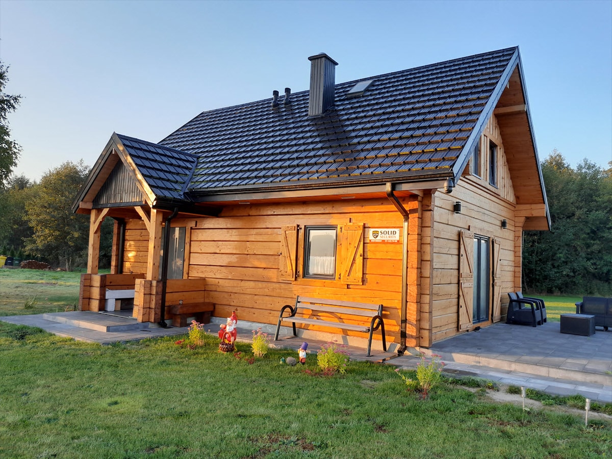 The exterior of a wooden house constructed from traditional fir logs is depicted. The house features a gabled roof, multiple windows, and a welcoming porch with seating. A manicured lawn surrounds the structure, with nearby greenery offering a natural backdrop.