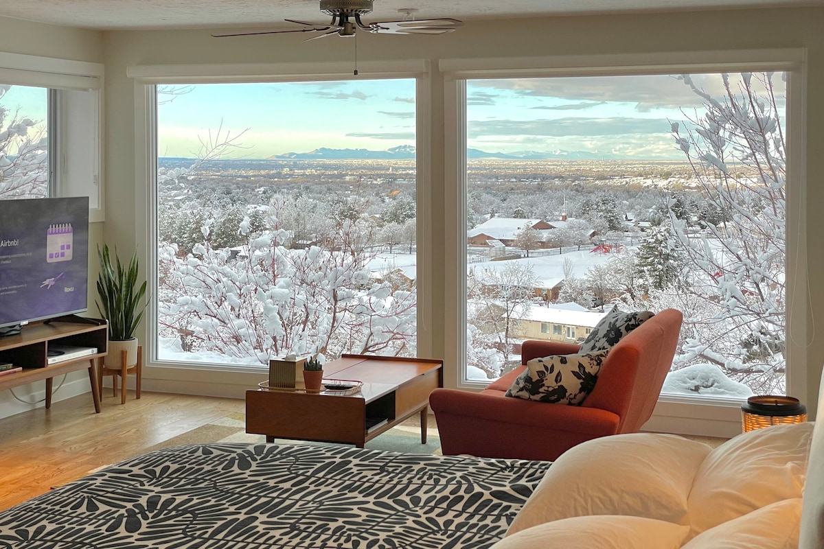 A spacious living area features a large window framing scenic snowy vistas of the surrounding landscape. A cozy red chair and a coffee table are positioned in front of the window, while a TV is visible on a wooden console. Soft bedding is neatly arranged on a bed in the foreground.