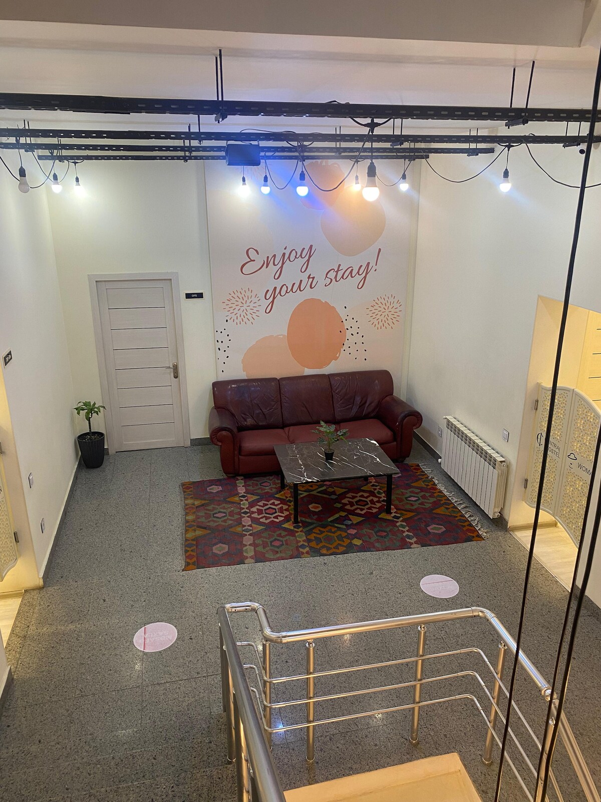 A welcoming common area is shown, featuring a comfortable red sofa and a decorative coffee table. Colorful rugs are placed on a smooth, gray floor. Wall art conveys a warm message, while soft lighting creates a cozy atmosphere. A small plant adds a touch of greenery.