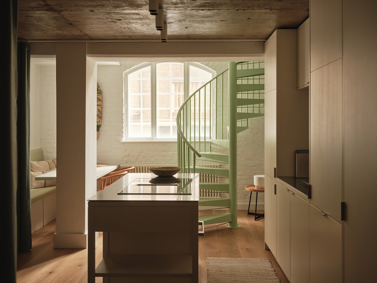 A spacious living area is depicted with a light color palette. A spiral staircase in mint green ascends to the second level. Natural light floods through large windows, illuminating the engineered wood flooring and a custom kitchen area. A comfortable sofa is positioned nearby.