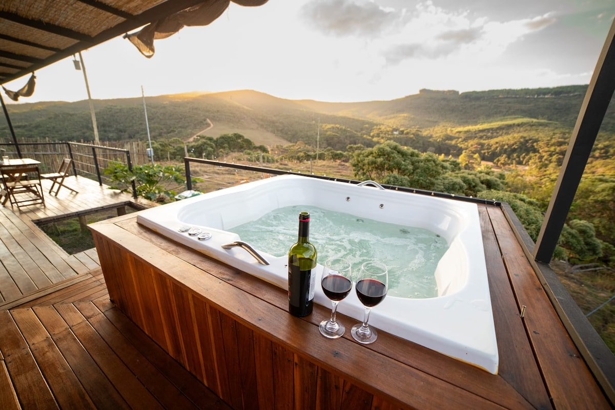A private hot tub is situated on a wooden deck, surrounded by views of rolling hills and lush greenery. Two glasses of red wine and a bottle are placed nearby, with soft evening light illuminating the tranquil scene.
