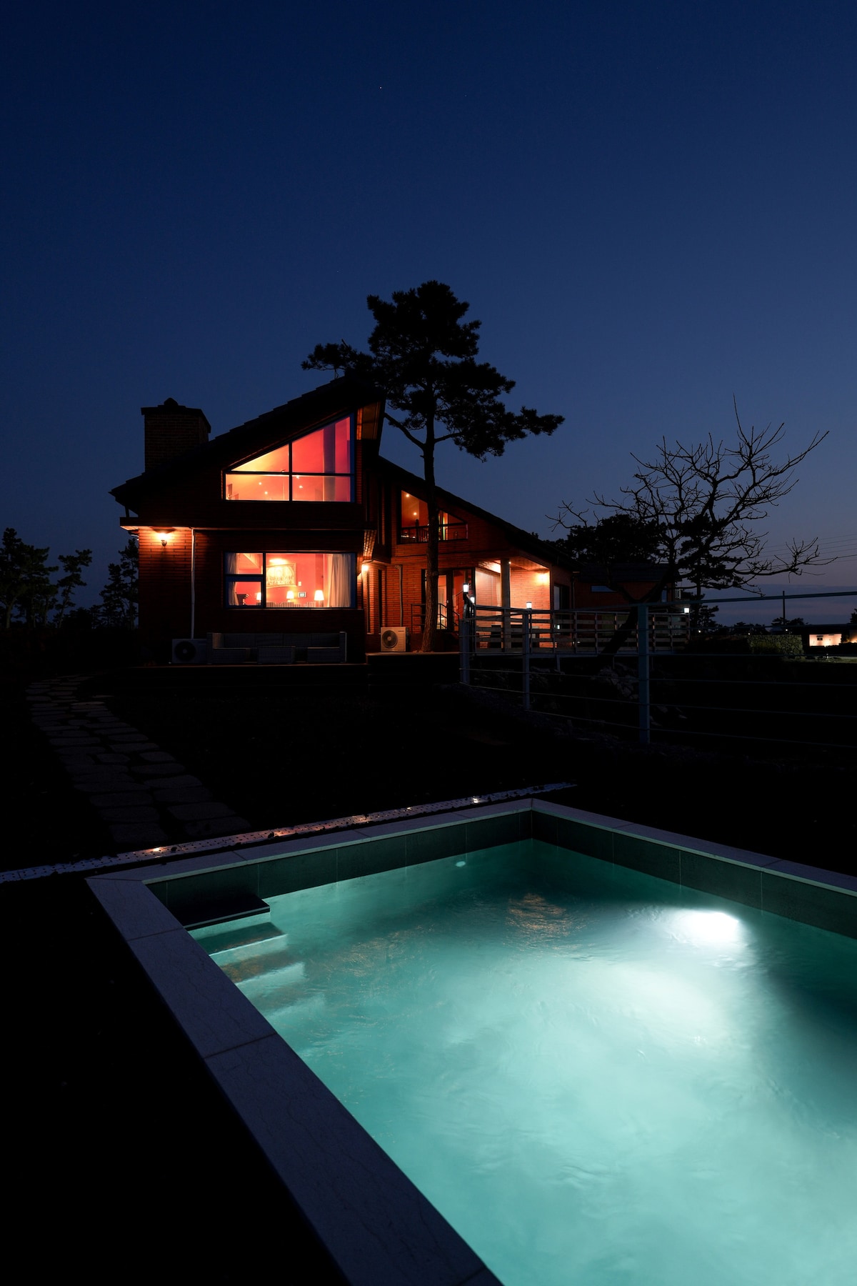 The exterior of a two-story wooden house is illuminated at night, showcasing large windows that emit a warm glow. A private swimming pool is visible in the foreground, featuring soft lighting that highlights its tranquil water.