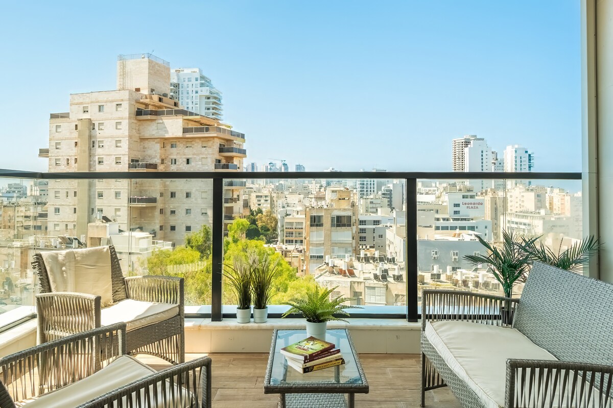 A balcony space offers a panoramic view of the city skyline, featuring modern buildings and greenery. Comfortable outdoor seating is arranged around a glass table, creating an inviting area for relaxation and enjoying the scenery.