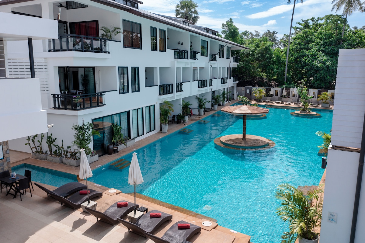 A spacious pool area is presented, featuring a large swimming pool with a central island and a shallow area for relaxation. Comfortable sun loungers and umbrellas line the poolside, surrounded by lush greenery and modern residential buildings.