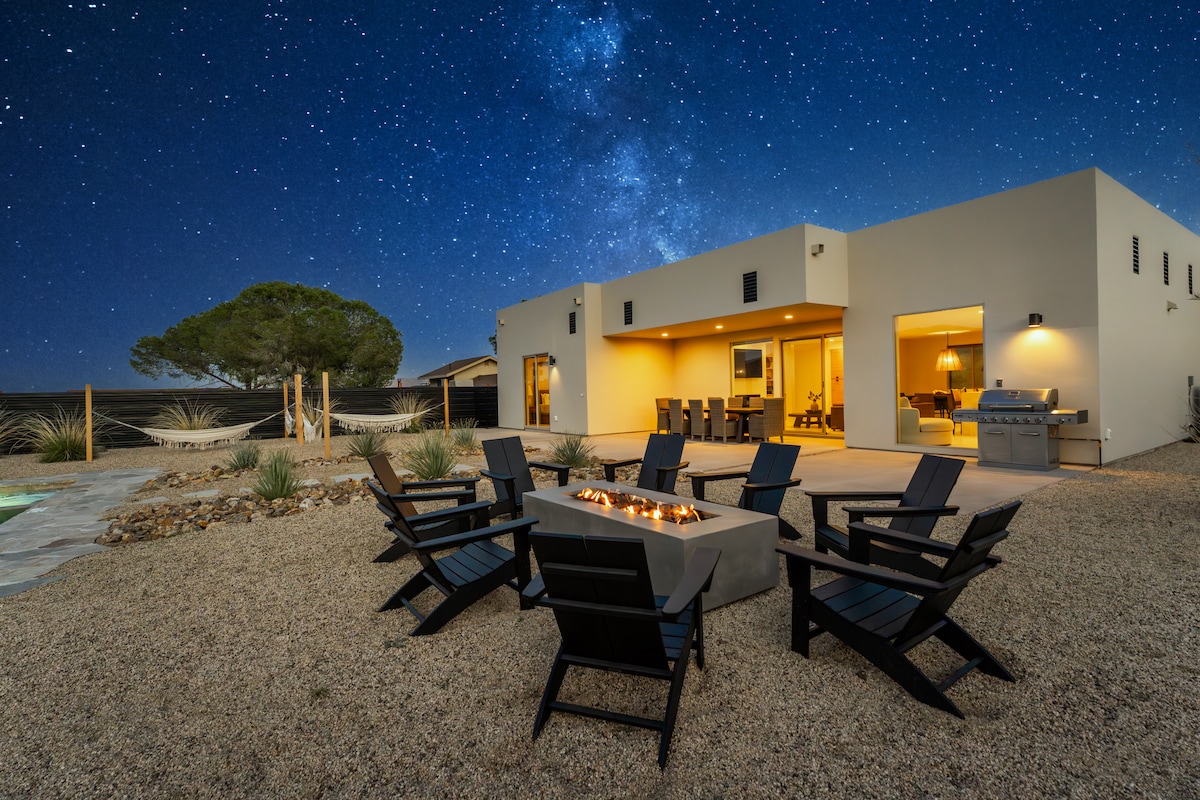 An outdoor gathering area is depicted, featuring a fire pit surrounded by six black wooden chairs. The backdrop showcases a modern home illuminated by warm light, set against a clear night sky filled with stars, emphasizing a tranquil desert oasis.